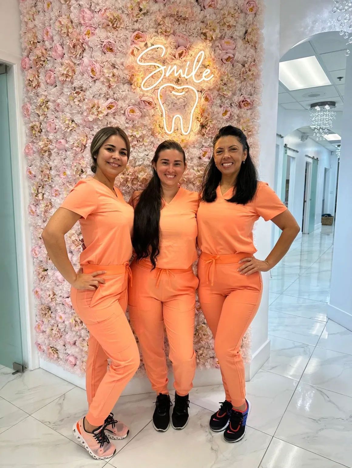 Three dental staff in peach scrubs pose by a flower wall and neon 