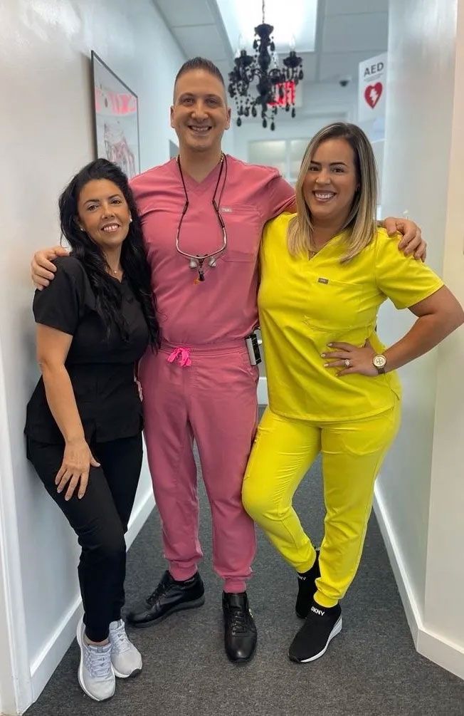 Three people in scrubs pose in a hallway. The center person wears pink scrubs, the other two wear black and yellow.