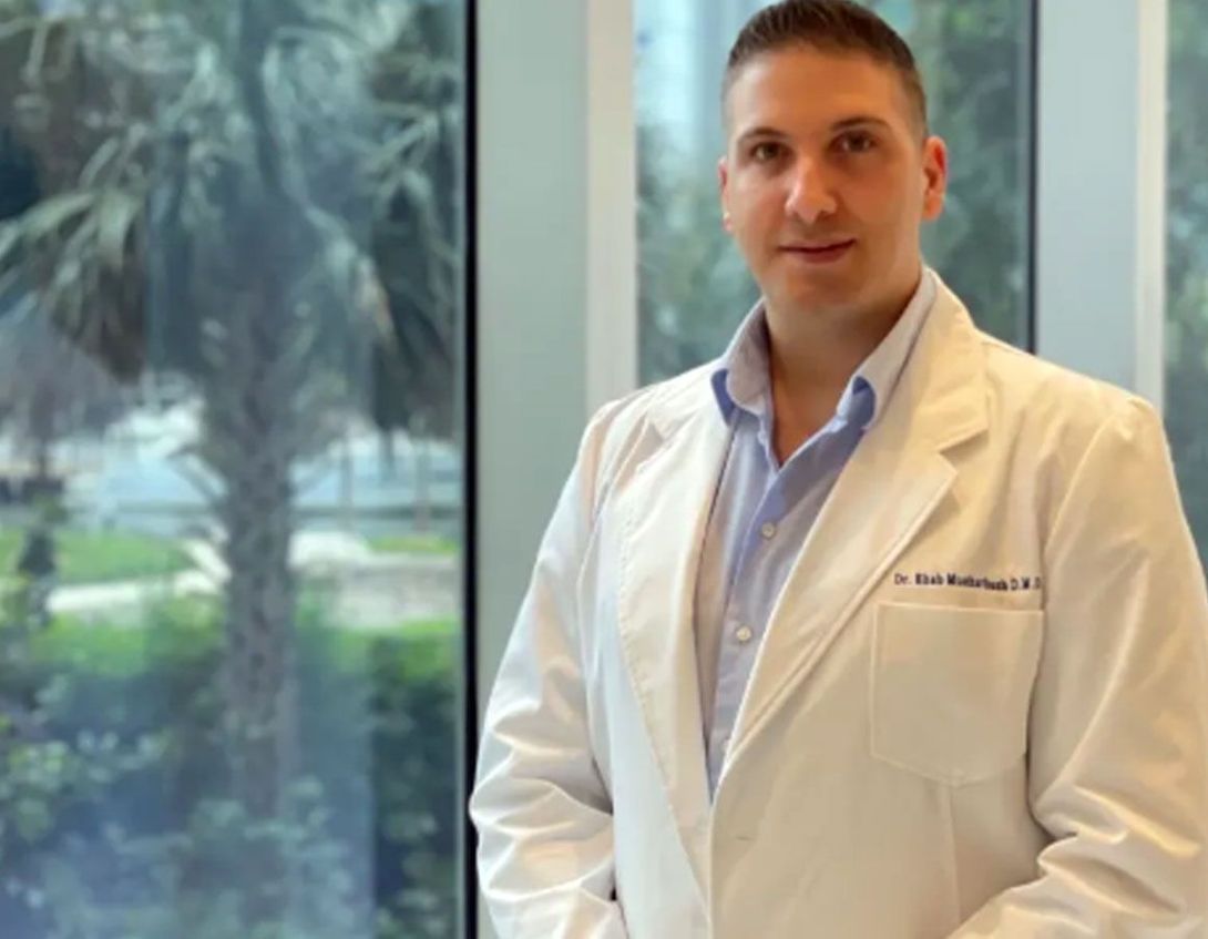 Man in white coat, light blue shirt, in front of a window. Palm tree visible outside.