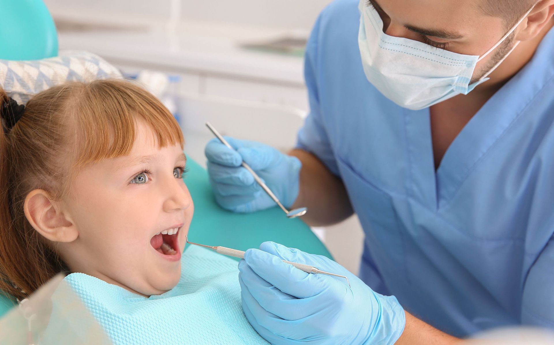 Child in dentist chair having teeth examined. Dentist in blue scrubs and mask.