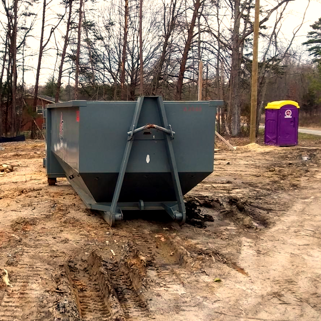 A dumpster is sitting in the dirt next to a purple portable toilet.