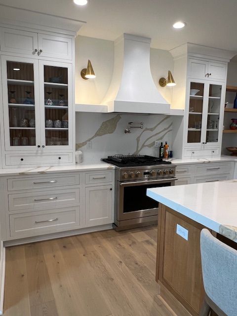 A white, modern kitchen featuring glass-front cabinets, a stainless steel range, and light wood flooring.
