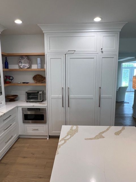 Modern kitchen with white panel-front appliances, a built-in microwave, open wooden shelves, and a marble-topped island.