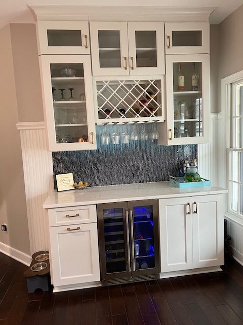A white kitchen bar unit with glass-front upper cabinets, a wine rack, under-counter wine cooler, and dark tile backsplash.
