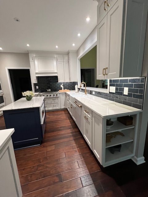 A modern kitchen featuring white cabinets, dark blue island, white countertops, dark gray backsplash, and wood flooring.