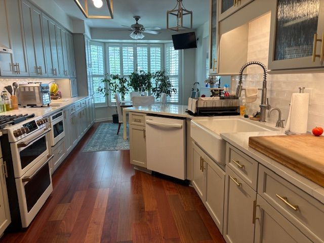 A galley-style kitchen with light gray cabinets, wood floors, a white farmhouse sink, and a sunny breakfast nook.