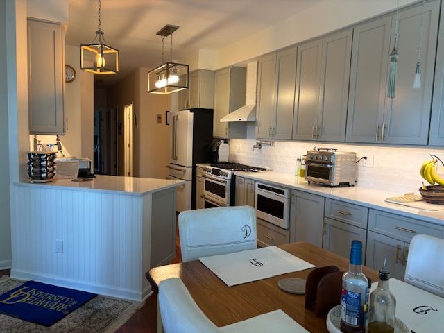 A modern kitchen with grey cabinetry, a white countertop, two pendant lights, and a dining table in the foreground.