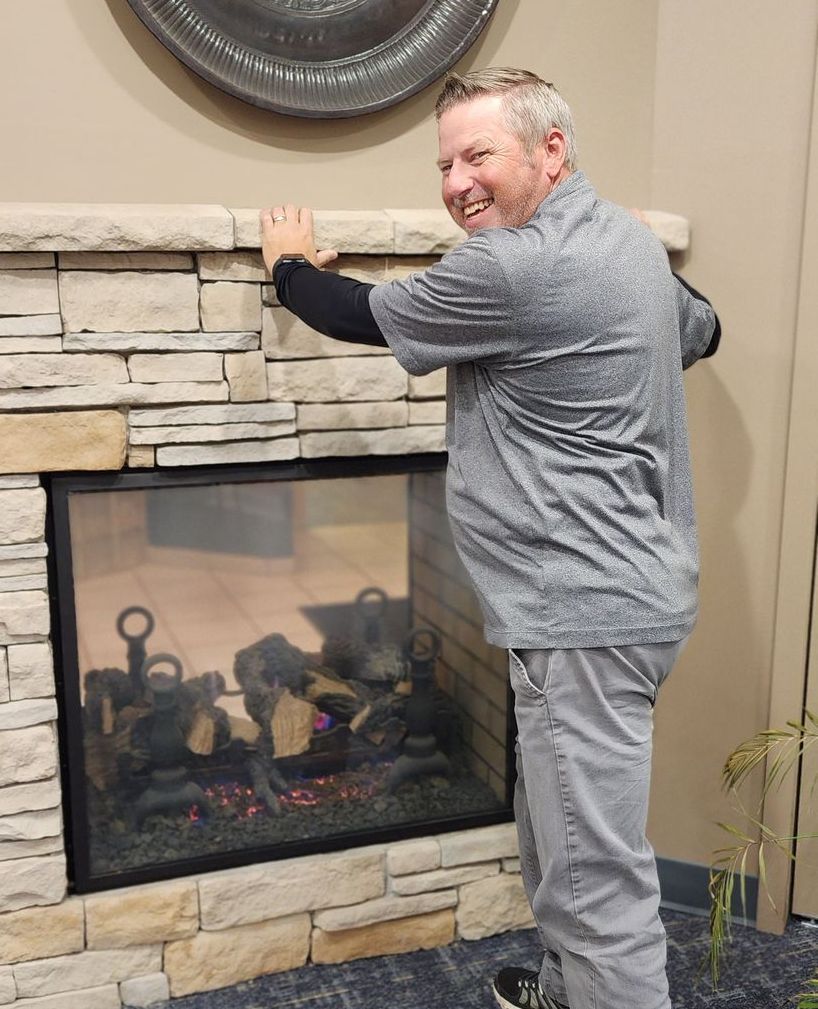 A man in a grey shirt is standing in front of a fireplace.