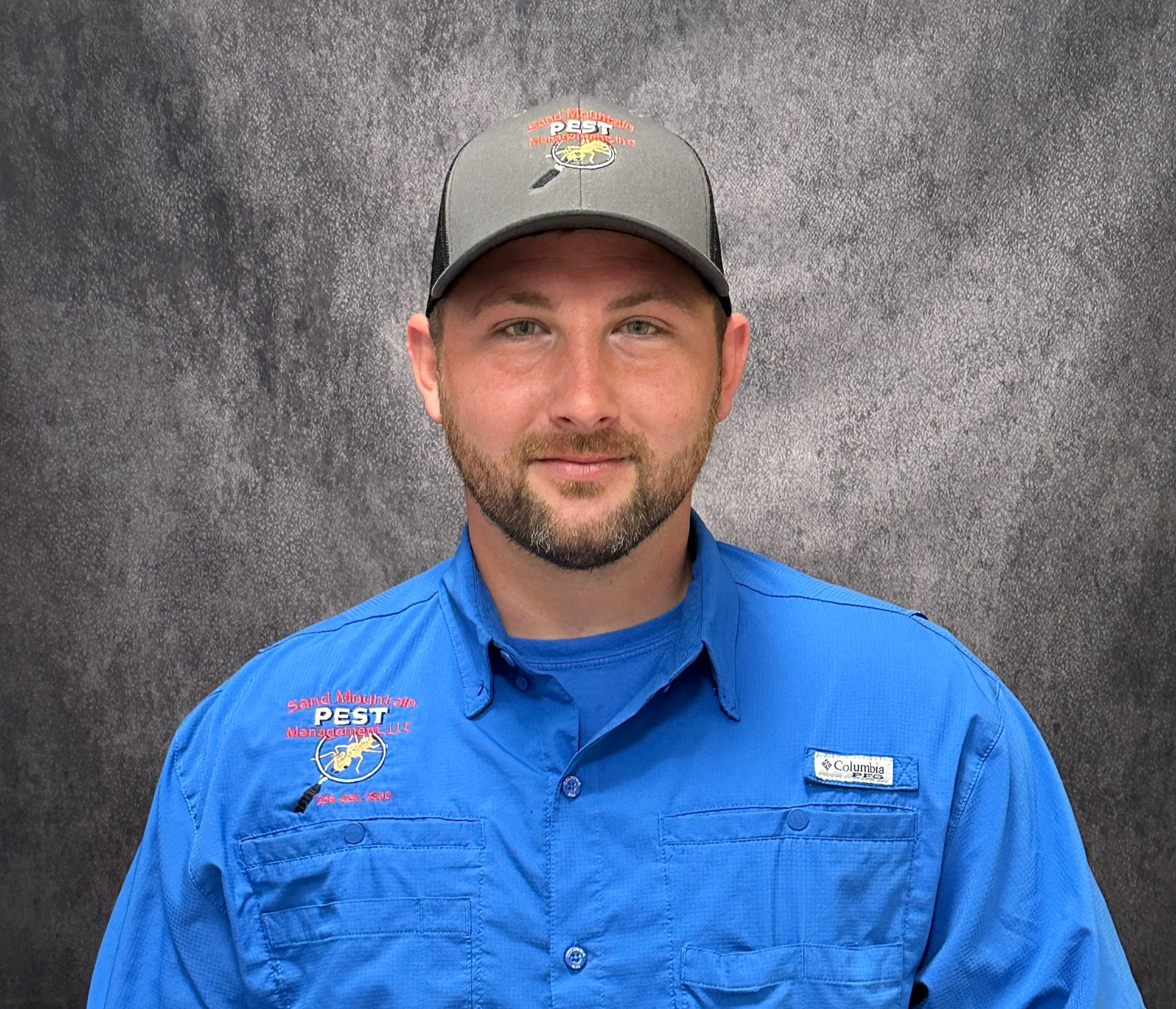 Man with brown hair and beard, wearing a black shirt in front of a brick wall.