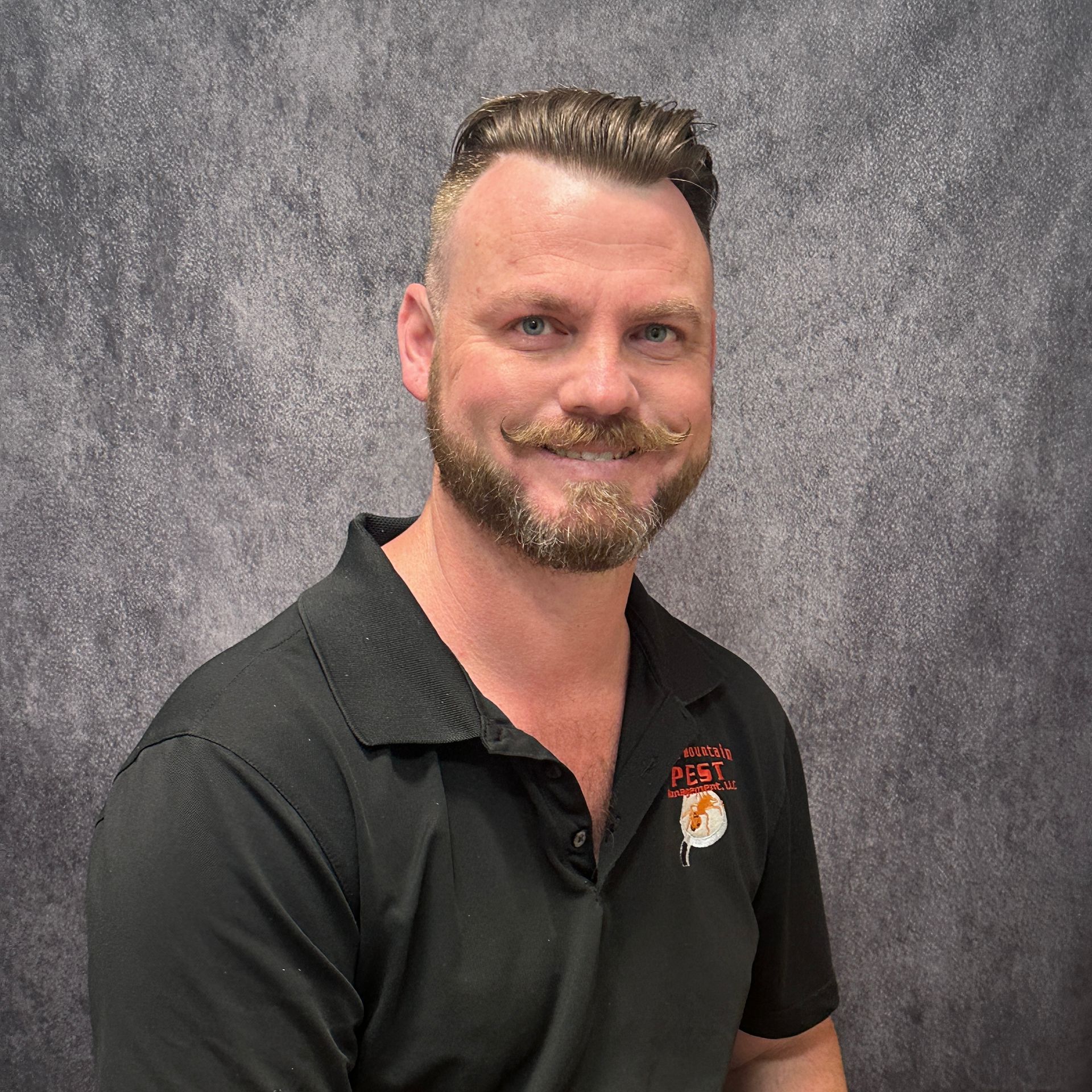 Man with a beard smiling in front of a brick wall. He wears a black collared shirt.