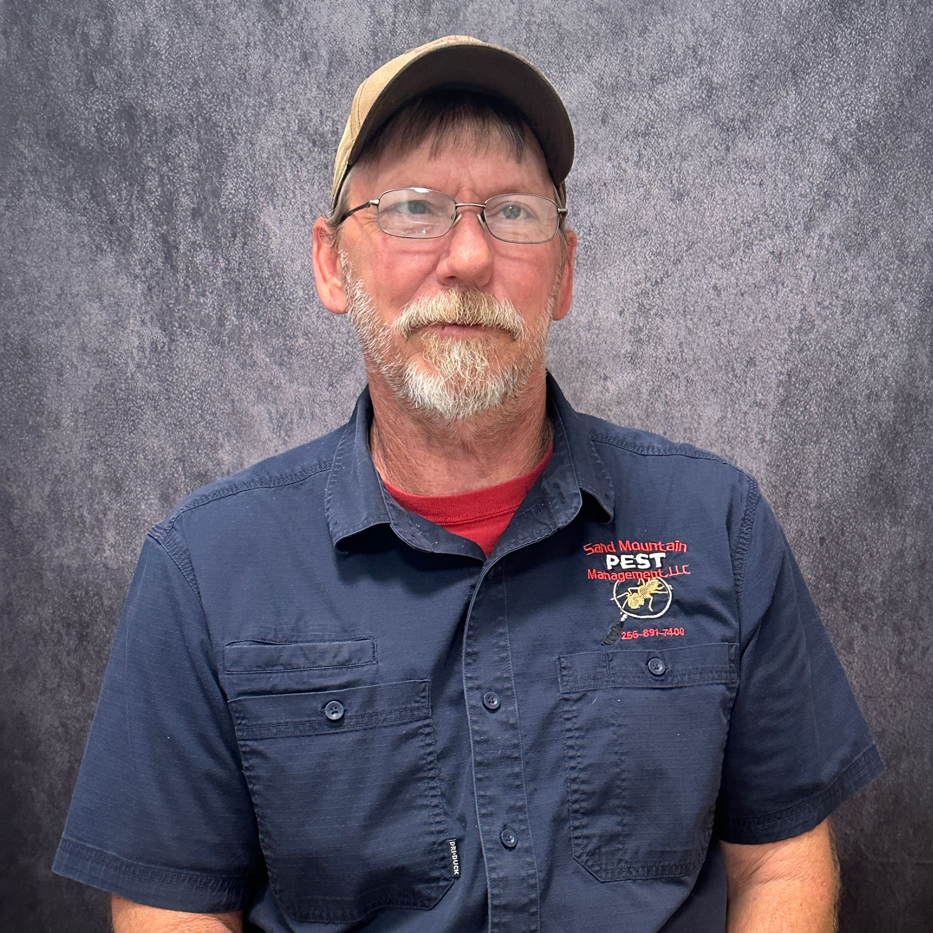 Man with glasses and a goatee, wearing a cap and black shirt, in front of a brick wall.