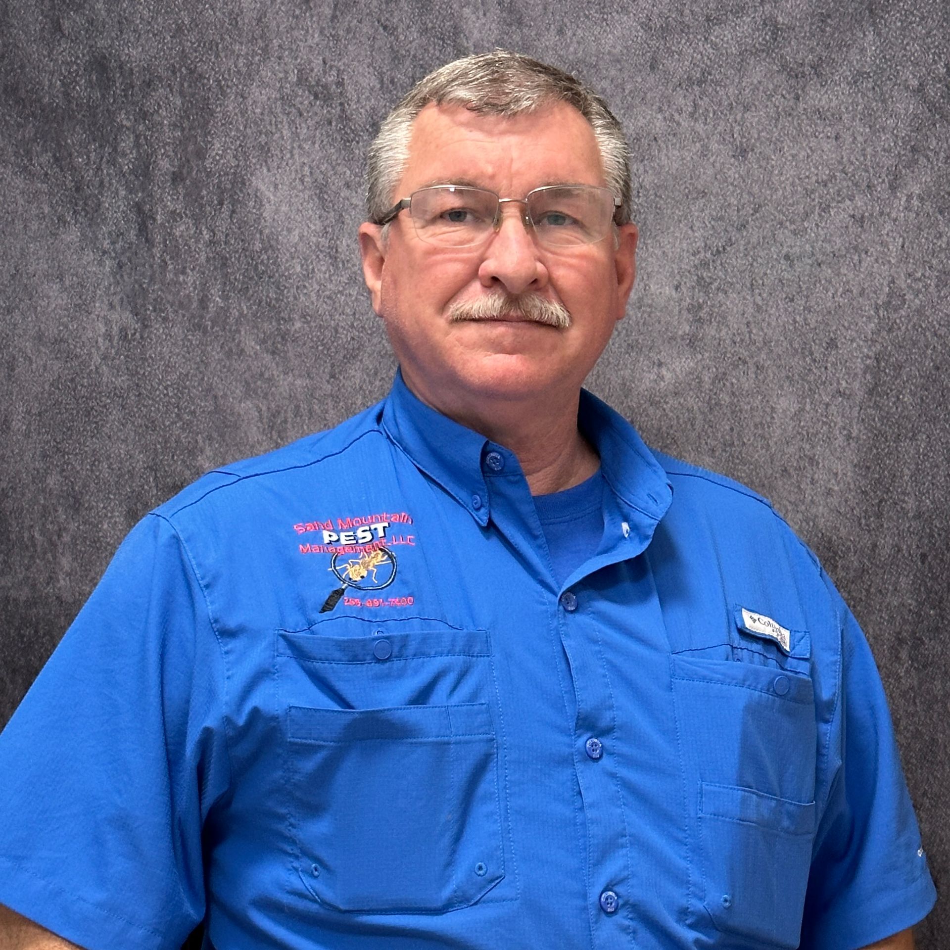 Man with glasses and grey hair in front of a brick wall, wearing a black shirt with a logo.