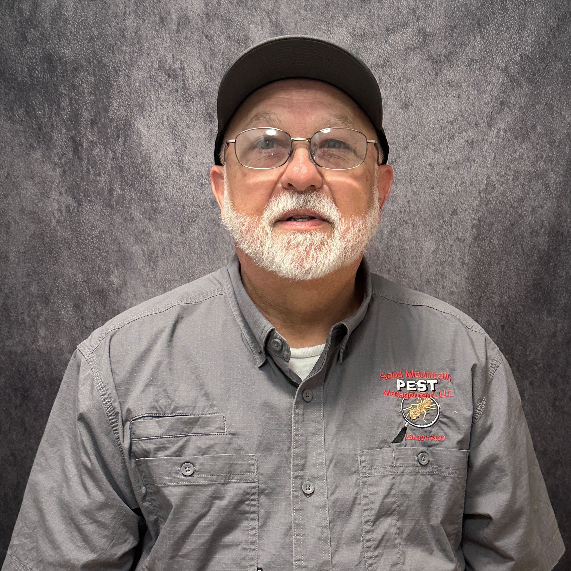 Man with glasses and white beard wearing a baseball cap and black shirt, in front of a brick wall.