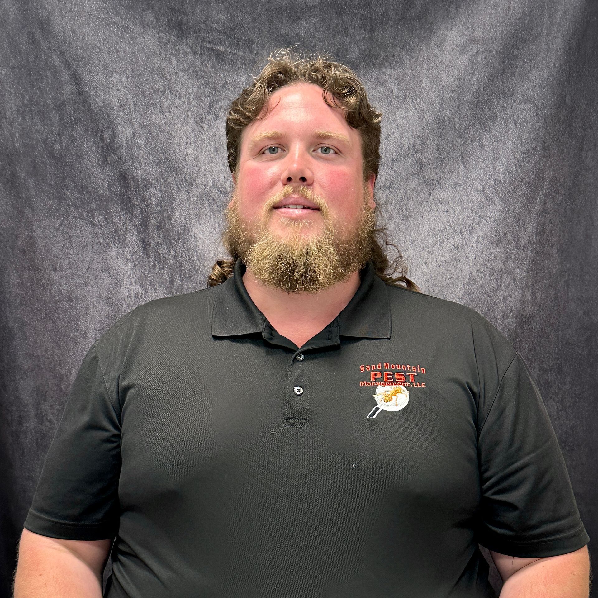 Man with a beard, mullet, and black shirt smiling in front of a brick wall.