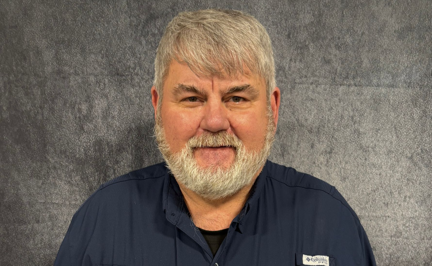 Man with graying hair and mustache stands in front of a brick wall, wearing a polo shirt with a company logo.