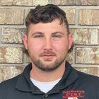 Man with brown hair and beard, wearing a black shirt in front of a brick wall.