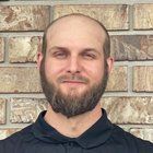 Man with beard and bald head smiles in front of a brick wall, wearing a dark shirt.