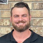 Man with a beard smiling in front of a brick wall. He wears a black collared shirt.