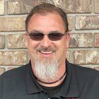 Man with a goatee and sunglasses smiling in front of a brick wall.
