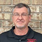 Man with glasses and grey hair in front of a brick wall, wearing a black shirt with a logo.