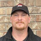 Man in black cap and jacket, ginger goatee, smiling in front of a brick wall.