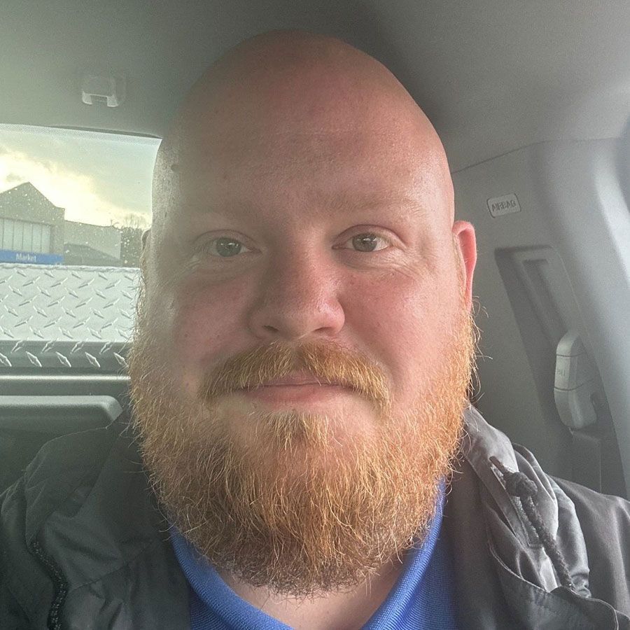 Bald man with a red beard in a vehicle. He's wearing a blue shirt and looking at the camera.