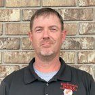 Man in black polo shirt stands in front of a brick wall. The shirt has a pest control logo.