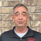 Man in black polo shirt stands in front of a brick wall.