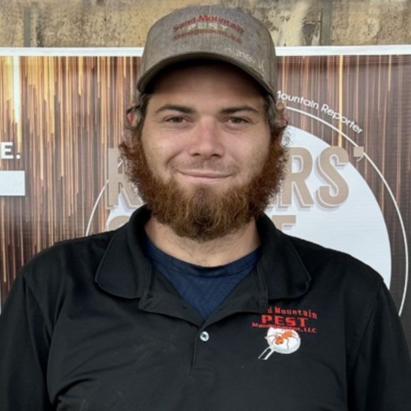 Man with beard and hat, wearing a black shirt, smiles in front of a sign.