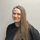 Woman with long gray hair smiles, wearing a black top against a plain wall.