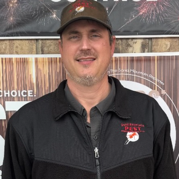 Man wearing a brown cap and black jacket smiles in front of a sign.