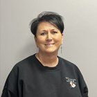 Woman with short dark hair smiles, wearing a black sweatshirt, standing against a gray wall.