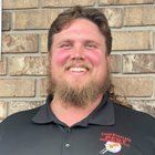 Man with a beard, mullet, and black shirt smiling in front of a brick wall.