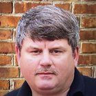 Man with salt and pepper hair and mustache stands in front of a brick wall.
