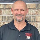 Man with a goatee smiling in front of a brick wall, wearing a black shirt with a company logo.