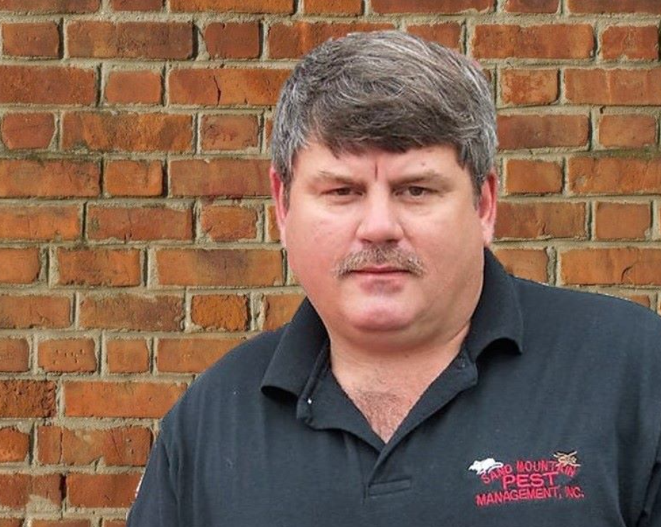 Man with graying hair and mustache stands in front of a brick wall, wearing a polo shirt with a company logo.