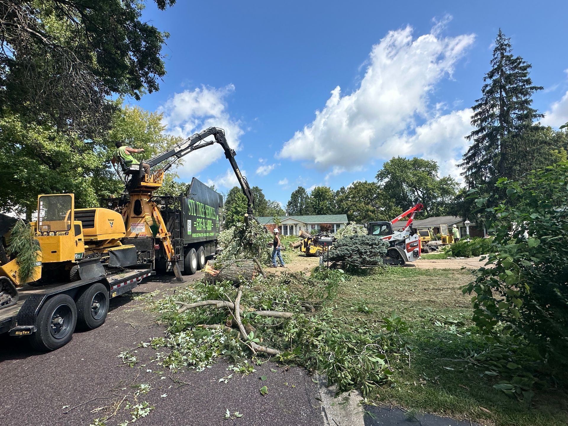 Tree removal: Large yellow chipper truck processing branches. Several other vehicles and people in yard.