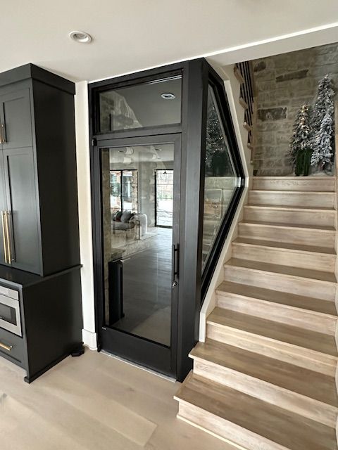 Modern black door with glass panel beside wooden stairs and stone wall.