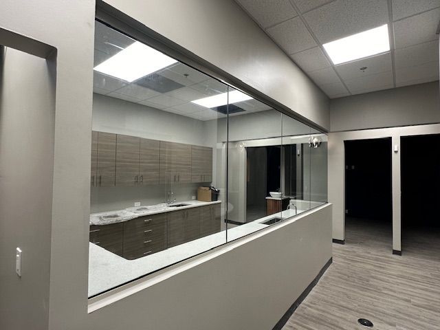 Hallway with a large glass window, looking into a medical office with beige cabinets and sink.