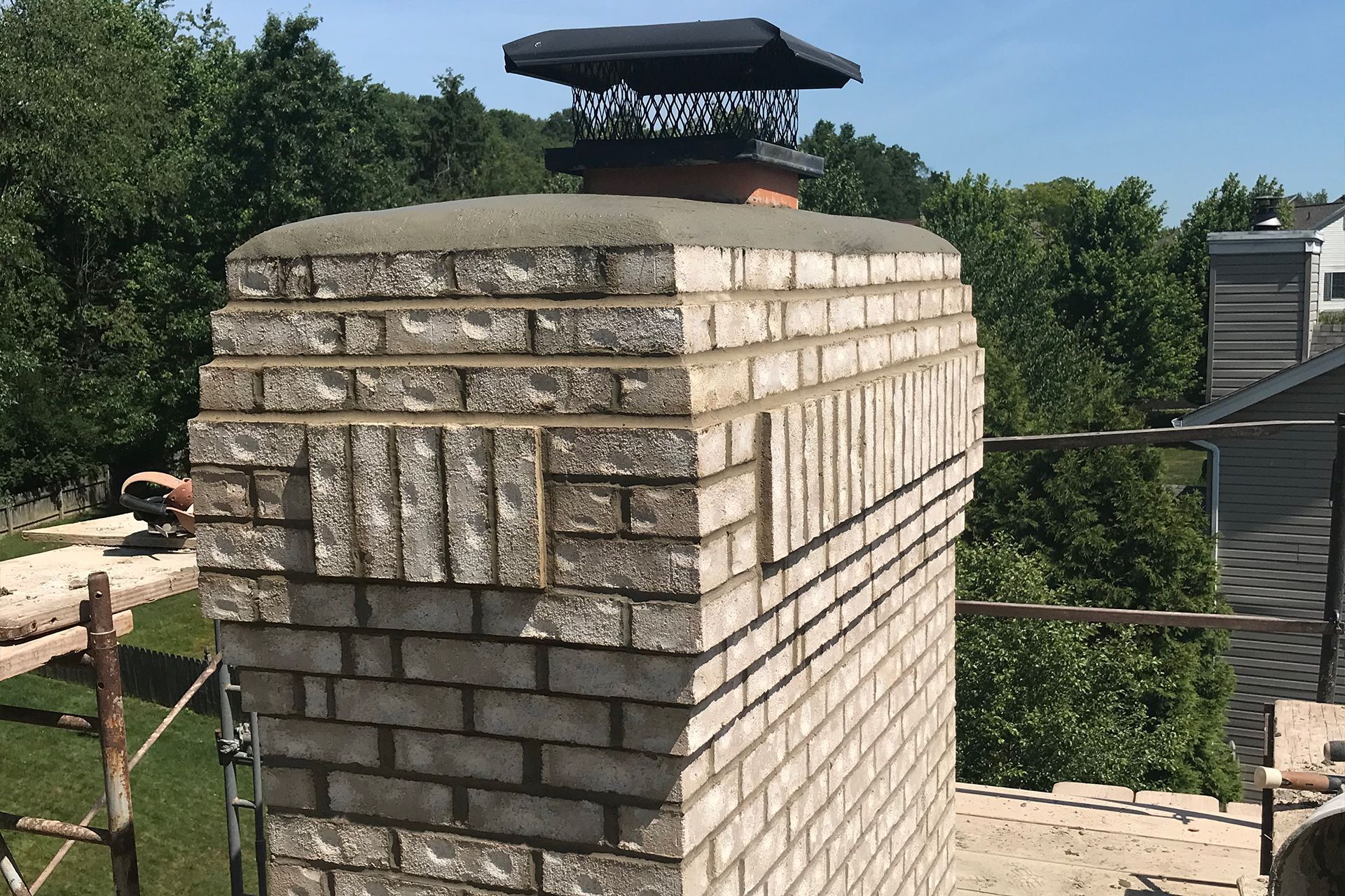 Brick chimney with cap against a backdrop of green trees and a blue sky.