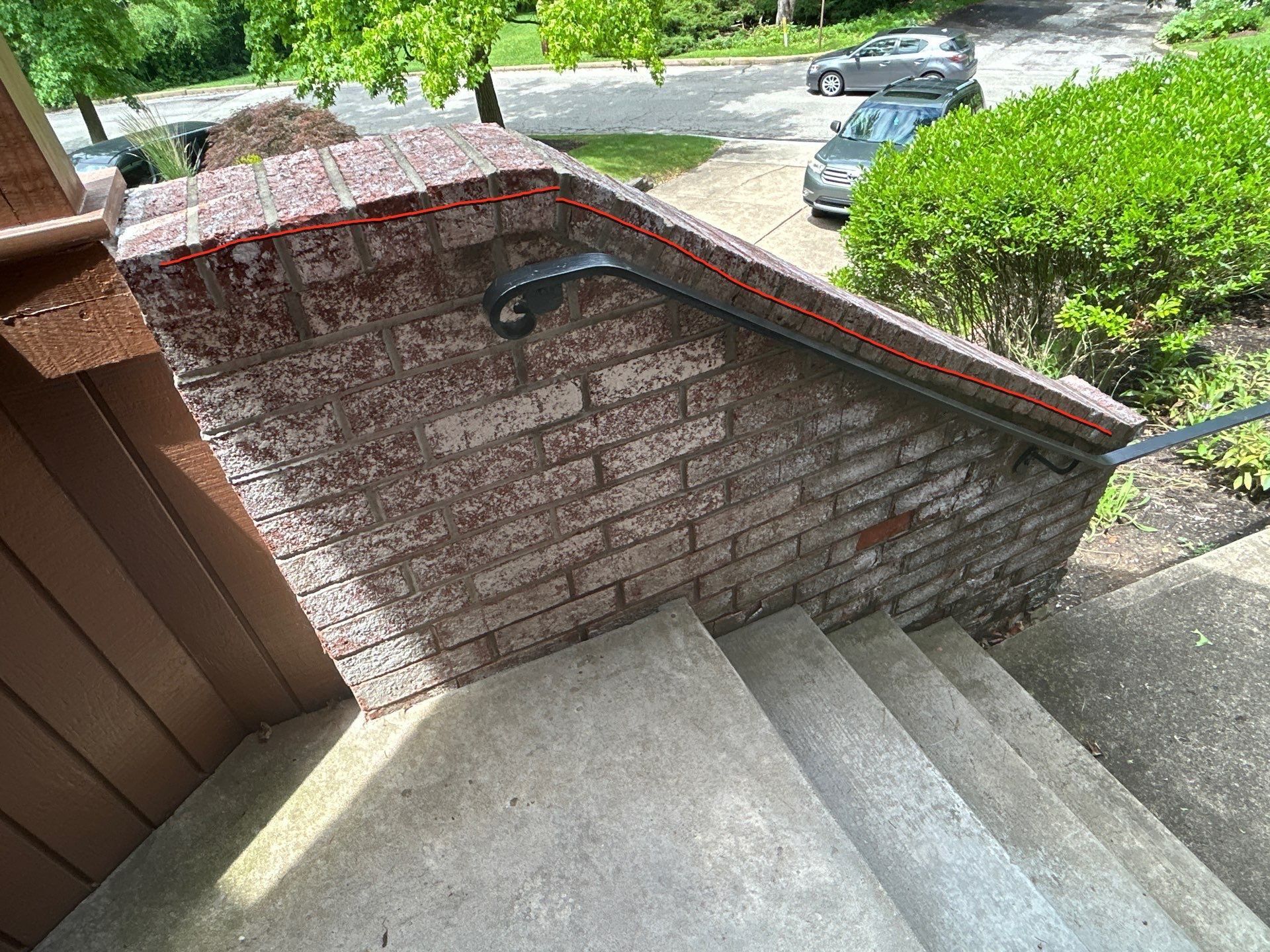 Brick railing with a black handrail, alongside outdoor concrete steps. Cars and greenery in the background.