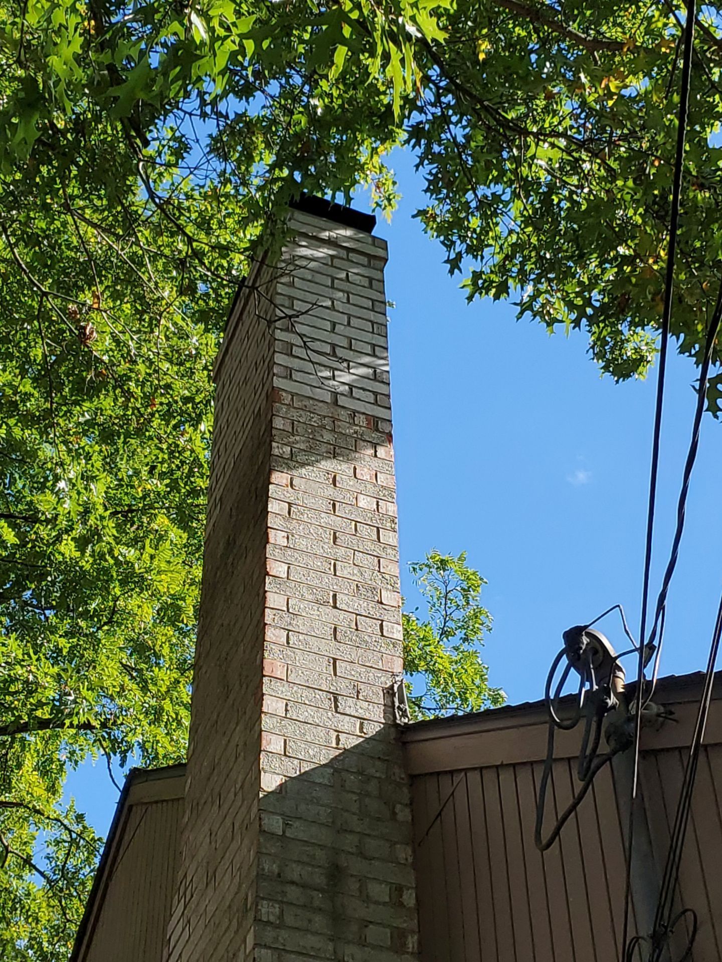 Brick chimney extending upwards against a blue sky, surrounded by green tree leaves.