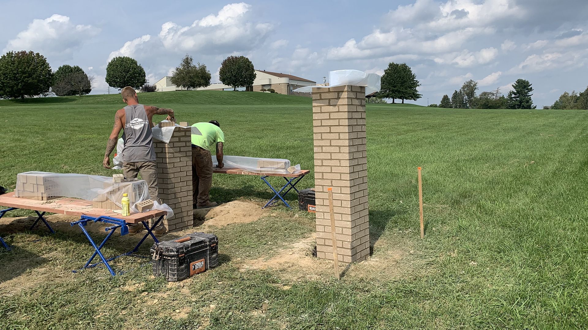 Two workers building brick columns in a field with a house visible in the distance.
