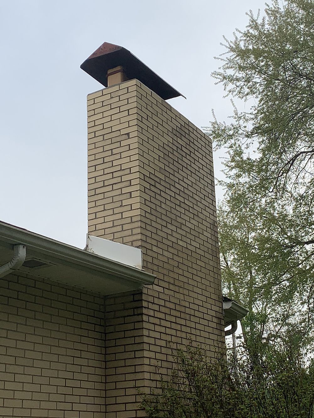 Brick chimney with a metal cap, attached to a light-colored brick house, under a cloudy sky.