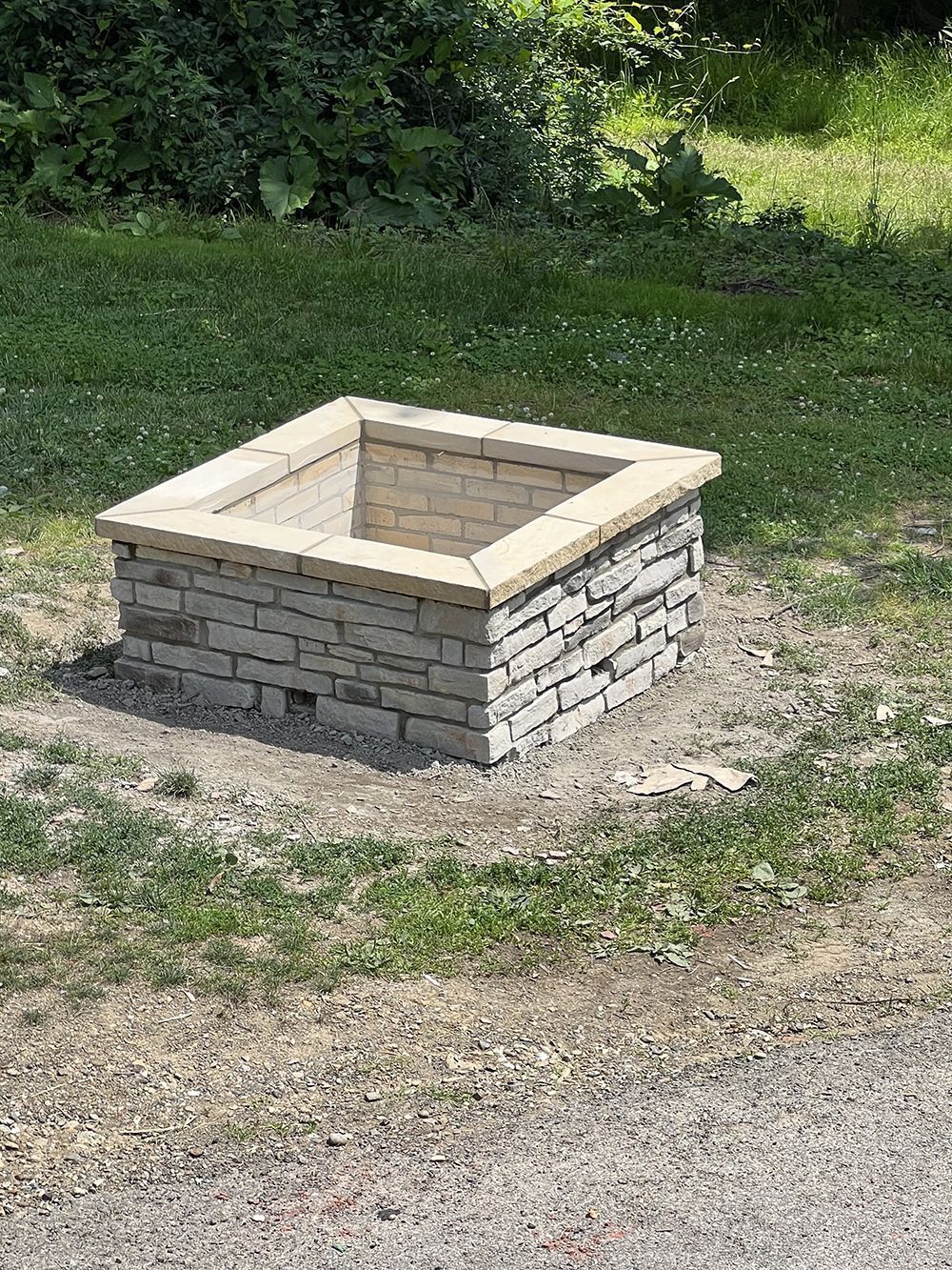 Square stone fire pit in a grassy area with surrounding foliage.