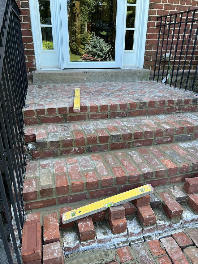 Brick steps being built, a spirit level rests on the bricks. Door and black iron railing are visible.