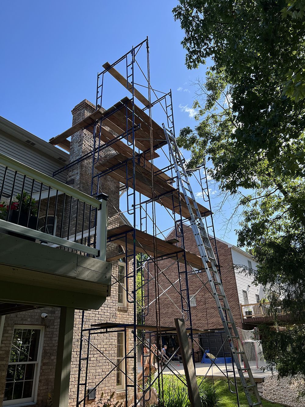 Scaffolding erected next to a brick chimney and house. Wooden platforms are visible. Blue sky in the background.