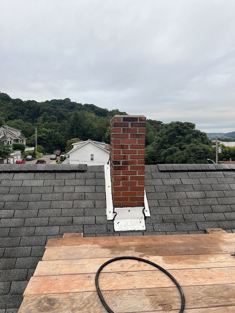 Brick chimney on a gray shingle roof, surrounded by white metal flashing. Overcast sky, trees in the background.