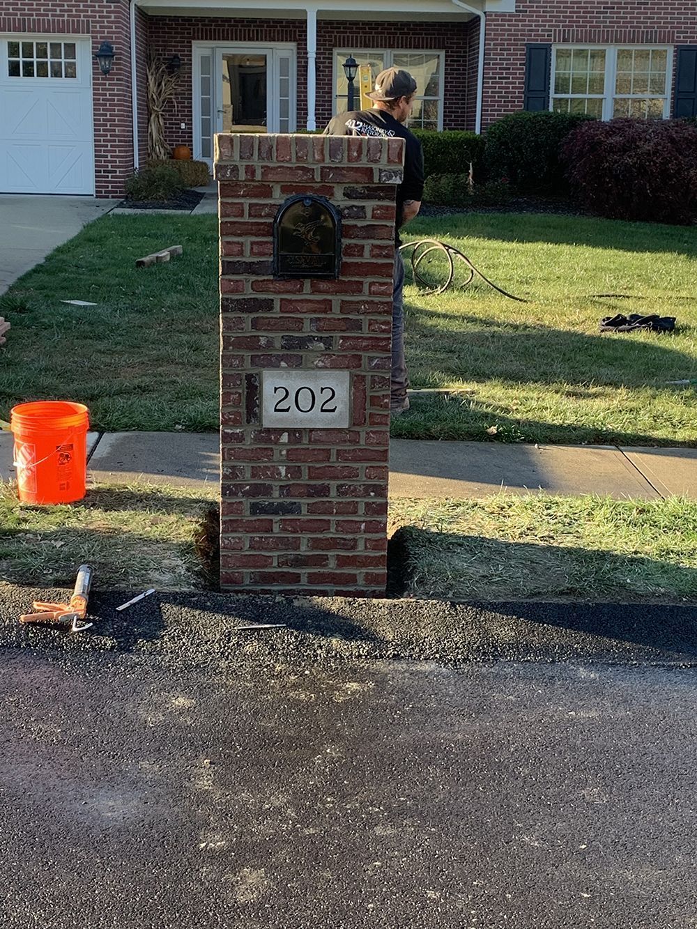 Brick mailbox with house number 202; person near it. Orange bucket, grass, asphalt.