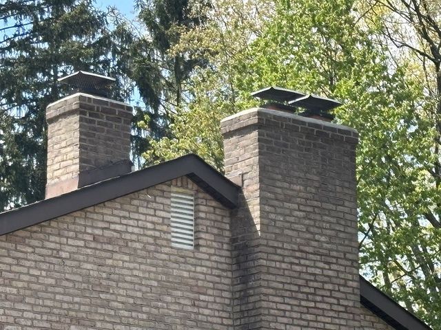 Brick chimneys on a roof with dark trim, topped with chimney caps, against a backdrop of trees.